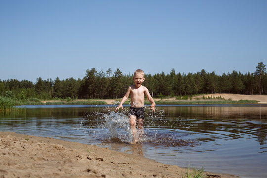 blond boy in black swimming trunks laughs and runs to the shore of the lake, water splashes from his feet.
lmage with selective focus