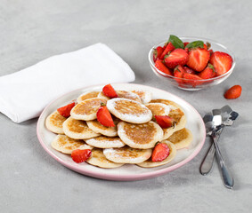 Mini pancakes with strawberries and powdered sugar on a plate on a light background