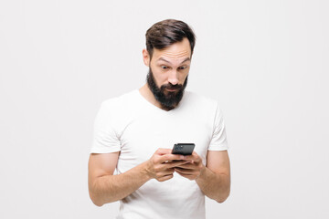Expressive guy in a white T-shirt plays video games on a smartphone, looks at the screen and reacts emotionally. Young man plays a game on the phone, the focus is on a smartphone in his hands