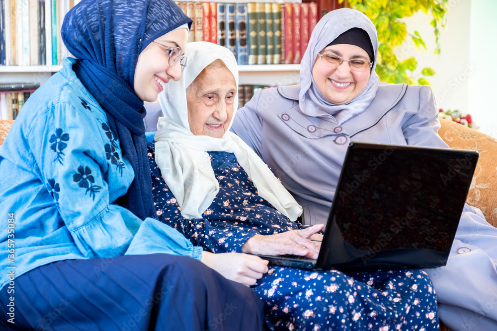 Wall mural happy arabic muslim grandmother and grand daughter and mother sitting togther on couch using technol