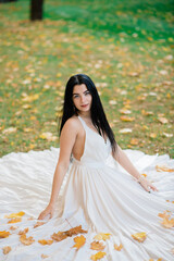 Young beautiful brunette female in a long white dress in autumn park.
