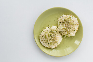 Two round grain toasts with fresh green alfalfa seed sprouts and cheese on a green plate with white background from above. Healthy food snack concept.