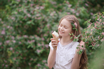 a girl with blond hair eats a delicious ice cream in a waffle cone, amid pink flowering bushes and greens, licks an ice cream