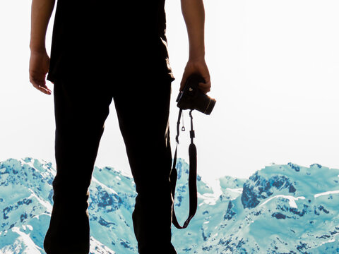 hombre joven con polera y pantalones negros con c&aacute;mara en la mano, de fondo monta&ntilde;as nevadas 