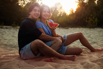 Young Caucasian woman and man eat watermelon on the beach at sunset.