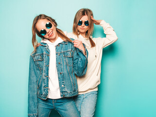 Two young beautiful brunette smiling hipster girls in trendy summer hoodie and jeans jacket clothes. Sexy carefree women posing near blue wall. Trendy and positive models having fun in sunglasses