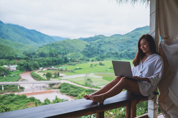 Fototapeta premium A beautiful asian woman working and typing on laptop computer while sitting on balcony with mountains and green nature background