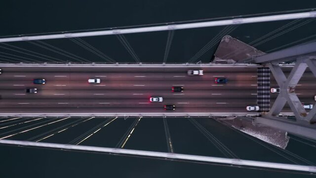 4K aerial overhead horizontal video of vehicles driving by the Bay Bridge towards San Francisco. Modern suspension bridge above the dark green bay waters is illuminated in the dark, California, USA