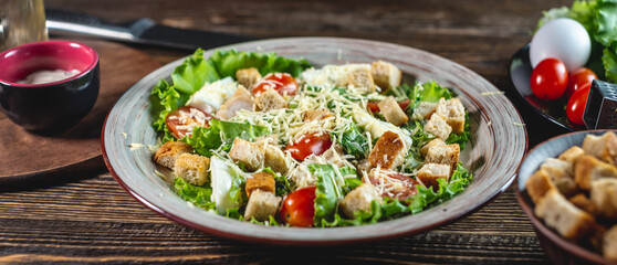 Plate with appetizing fresh Caesar salad on a wooden table and ingredients for cooking it. Top view