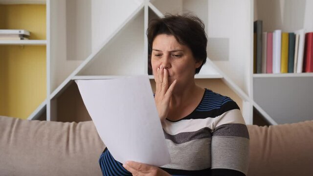 Sad woman sitting on couch at home reads received bad news holds documents paper letter feels desperate about financial problems. expulsion concept