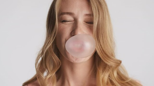 Beautiful funny blond girl joyfully blowing bubblegum over gray expression. Happy expression