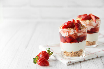 Homemade layered dessert with fresh strawberries, cream cheese or yogurt, granola and strawberry jam in glasses on white wood background. Healthy organic breakfast or snack concept. Selective focus.