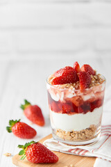 Homemade layered dessert with fresh strawberries, cream cheese or yogurt, granola and strawberry jam in glass on white wood background. Healthy organic breakfast or snack concept. Selective focus.