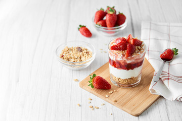 Homemade layered dessert with fresh strawberries, cream cheese or yogurt, granola and strawberry jam in glass on white wood background. Healthy organic breakfast or snack concept. Selective focus.