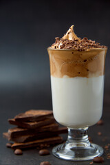 Glass of trendy fluffy creamy dalgona or whipped instant coffee with milk, coffee beans and chocolate on the dark background. New popular food and drink trend concept. Copy space.