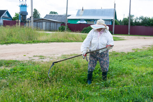 A woman mows fresh green grass with a scythe on a cloudy summer day.
