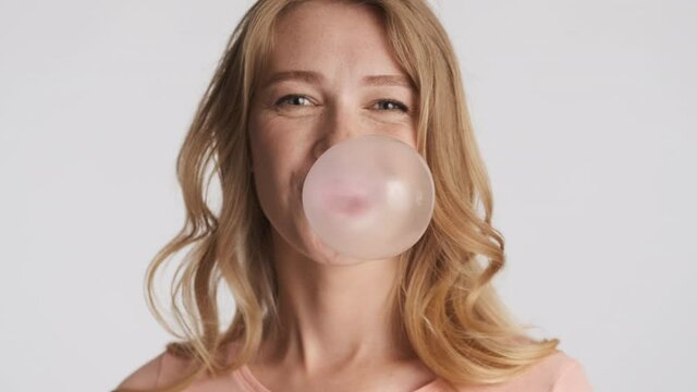 Young pretty funny blond woman happily blowing bubblegum over gray background