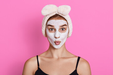 Young woman with cream mask on her face and hair band on head, girl with rounded lips looking...