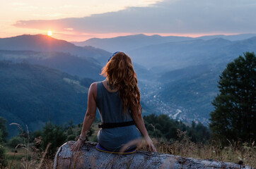 Naklejka na ściany i meble A girl sits on the edge of the cliff and looking at the sun valley and mountains. Woman sitting on mountain top and contemplating the sunset. Sunset in Ukrainian Carpathians