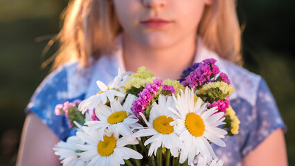 Obraz na płótnie Canvas Blonde girl holds a bouquet of flowers, stands in a meadow at sunset. Closeup shot