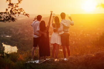 Company of young friends celebrating at picnic at sunset. They standing with glasses of white wine ad enjoying perfect sunset and landscape. Concept of friendship Shoot from back. 