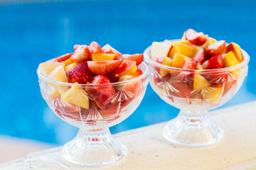 Two bowls of fresh fruit salad with swimming pool in background.