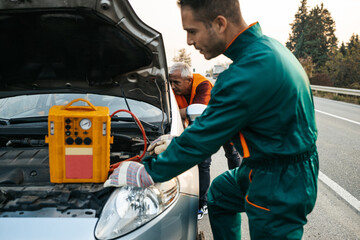 Two road assistant workers in towing service trying to start car engine with jump starter and energy station with air compressor. Roadside assistance concept.