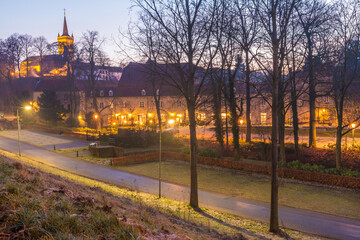 Rural road between leafless trees with Elsloo Castle illuminated with yellow lights from pole lamps in the background, calm and cold winter sunrise seen from a hill in South Limburg, Netherlands