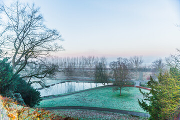 Leafless trees, green grass with ice, a small pond in Kasteelpark Elsloo seen from a hill, cold winter dawn with a light haze in South Limburg, the Netherlands Holland