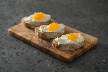 Three ciabatta slices with ricotta and orange marmalade on olive wood board on terrazzo surface