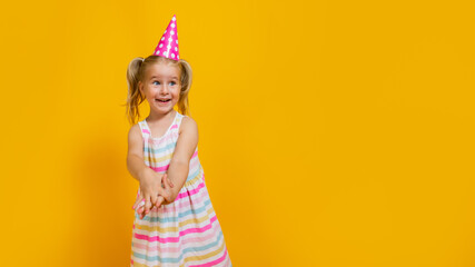 Happy birthday child girl with two ponytales in pink cap on colored yellow background. Copyspace.