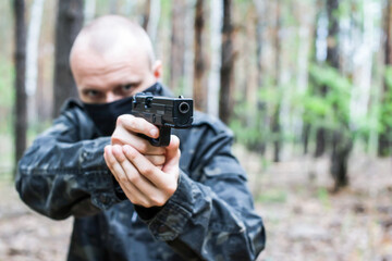 A man in dark military clothes and a mask is aiming from a weapon in the forest, close-up