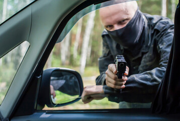 a man in camouflage and a mask with a pistol aims at the driver through the car window