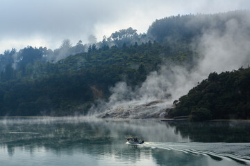 Orakei Korako geothermal area in the Taupo Volcanic Zone, New Zealand. Steam pours from a sinter terrace in the forest. In the foreground, a boat is crossing the Waikato River