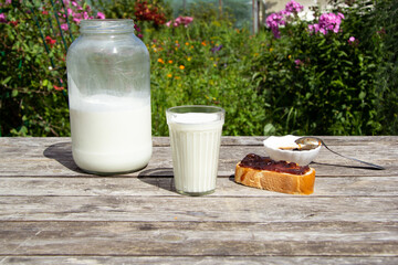 A faceted glass of milk stands on a wooden table. There is a large two-litre can of milk nearby. On the other side lies a loaf slice of jam