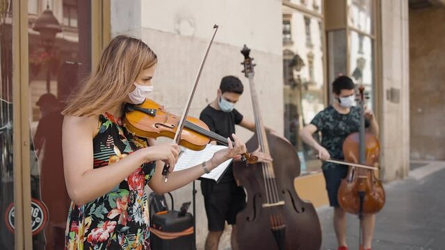 trio strings musical band wearing protective masks playing music outside in the street in pedestrian zone in pandemic time