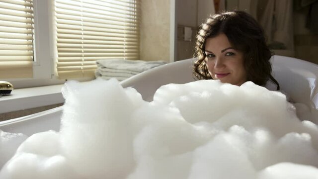 Young happy woman in the bath with white foam. Video. Portrait of a brunette female with curly hair and makeup on her face taking bubble bath at home.