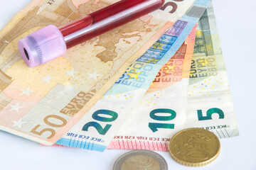 Close up of euro bill notes and coins with a tube full of blood on a white background. Coronavirus concept.