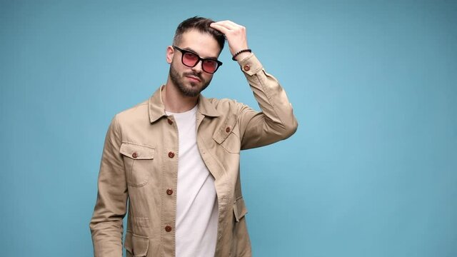 sexy young fashion man with sunglasses, running fingers through hair, adjusting coat and moving, arranging hair and moving in a side view position on blue background