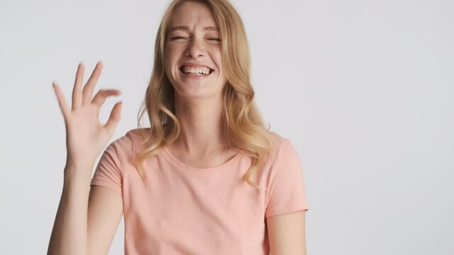 Young cheerful blond woman happily winking showing okay gesture and laughing on camera over gray background. Fooling around expression