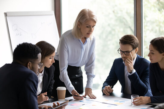 60s businesswoman project leader and diverse millennial workmates learn data of financial report during group meeting in boardroom. Do analysis and research, engagement in teamwork, brainstorm concept