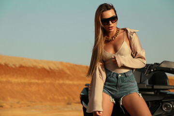 beautiful sexy woman with dark hair in casual clothes and accessories posing in desert with safari car - obrazy, fototapety, plakaty