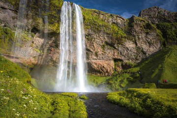 Waterfall Seljalandsfoss falling from high cliff in Iceland and tourists near it. The waterfall drops 60m and is part of the Seljalands River. Visitors can walk behind the falls into a small cave.
