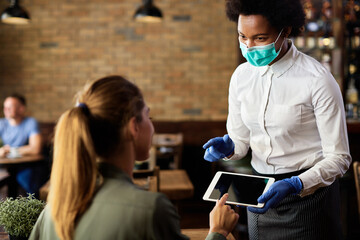 Black waitress wearing protective face mask while taking order on touchpad from her customer.