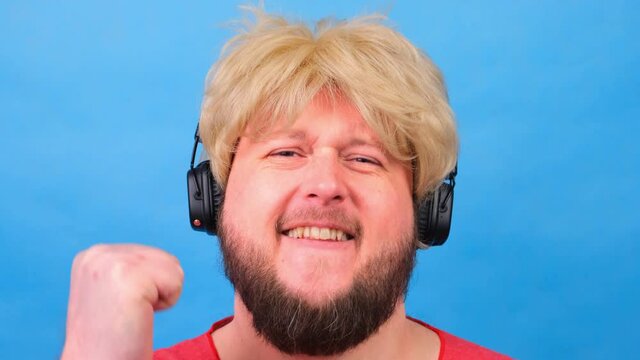 laughing freaky fat man in a wig and a pink T-shirt in his hands listens to music on headphones and dances on a blue background