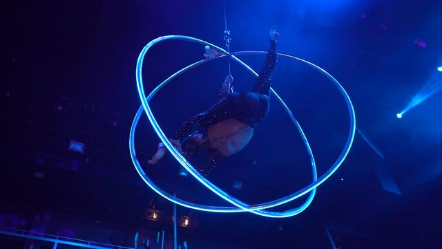 Flexible girl doing splits in aerial acrobatic hoop upside down illuminated in blue light, night club performance, gymnast performance