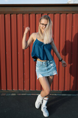 Beautiful millennial girl with a modern haircut and glasses posing cheerfully on a red wall. She wears a mini jeans skirt and easy summer blouse