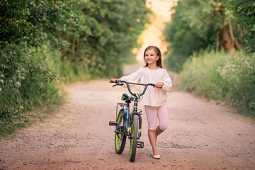 Fototapeta na wymiar little girl goes with a bicycle on a rural road in nature at sunset 