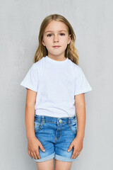 Little child girl in t-shirt, jeans shorts and sneakers posing near the wall