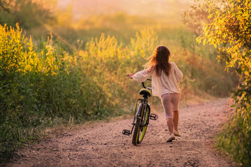 little girl goes with a bicycle on a rural road in nature at sunset	